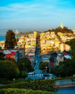 View down a San Francisco hillside street toward Coit Tower, the bay, and the Bay Bridge at golden hour, illustrating the Bay Area housing market in 2026.